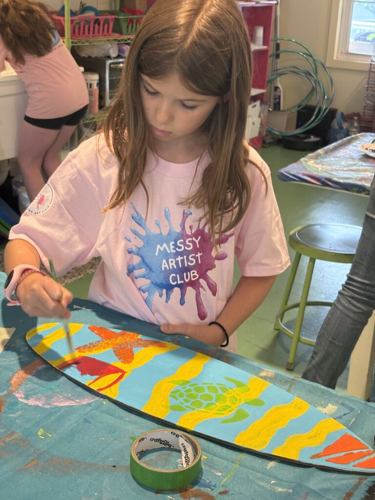 Young artist painting a colorful surfboard during Surfin’ USA Art Camp at Pink Art Pixie in Joppa, Maryland