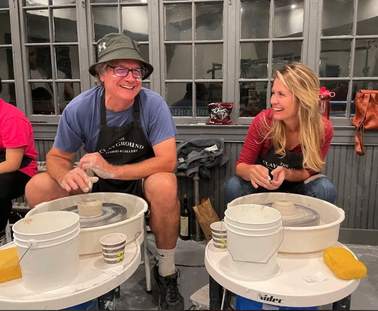 The Pixie and her Elf sit at pottery wheels, smiling and laughing while shaping clay. Both wear aprons; the man wears a hat and glasses. They appear to be enjoying a pottery class together in a cozy, well-lit studio. at Pink Art Pixie in Joppa, Maryland